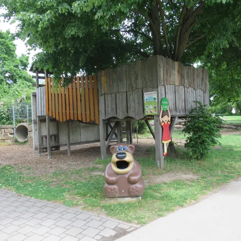 Ein Spielplatz mit einem Holzspielhaus und einer großen grünen Baumkrone. Vor dem Spielhaus steht eine bunte Figur eines Bären.