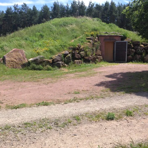 Ein kleiner Hügel mit einer Steinumrandung und einer Eingangstür. Der Platz ist von grünem Gras und Bäumen umgeben.