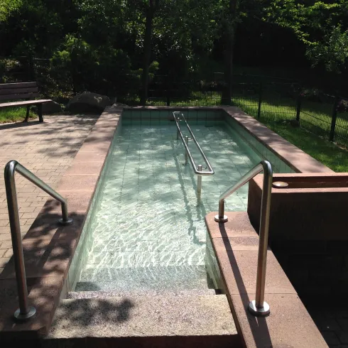 Ein privater Außenwhirlpool mit klarem Wasser und Metallgeländer. Im Hintergrund sind grüne Bäume und eine Parkbank sichtbar.