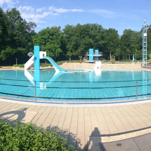 Ein Schwimmbad mit klarem Wasser und einem Sprungturm. Die Umgebung ist grün und sonnig.