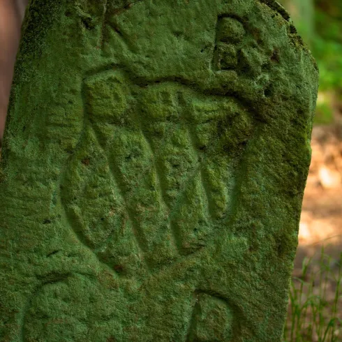Ein verwitterter Stein mit eingravierten Buchstaben und Symbolen. Die grüne Moosschicht deutet auf das Alter des Steins hin und verbindet ihn mit der Natur.