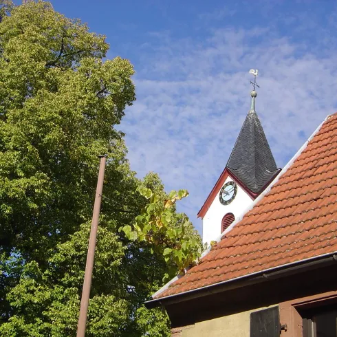 Ein hübsches Gebäude mit einem spitzen Dach und einer Uhr. Im Hintergrund ist ein großer, grüner Baum und ein blauer Himmel zu sehen.