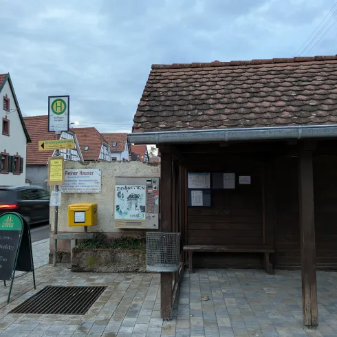 Eine Bushaltestelle mit einem überdachten Wartenbereich. Im Hintergrund sind Häuser und ein Parkplatz sichtbar.