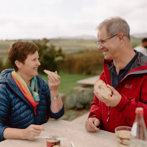 Ein lächelndes Paar genießt Essen im Freien. Im Hintergrund sind Weinberge und eine grüne Landschaft zu sehen.