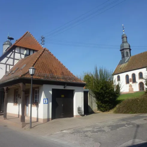 Ein malerisches Dorf mit einem traditionellen Fachwerkhaus und einer Kirche im Hintergrund. Der Himmel ist klar und blau.