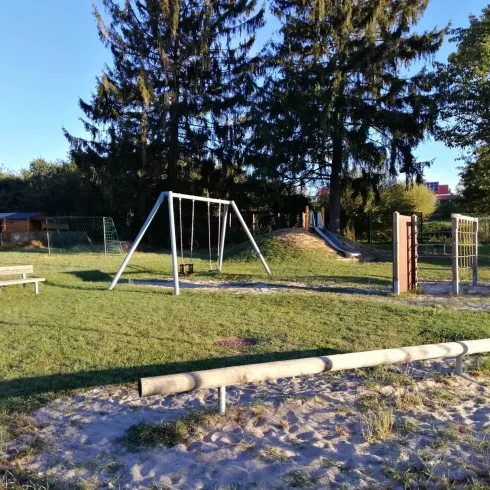 Ein Spielplatz mit einer Schaukel und einem Klettergerüst steht im Vordergrund. Umgeben von grünen Rasenflächen und hohen Bäumen bietet der Platz eine einladende Atmosphäre.