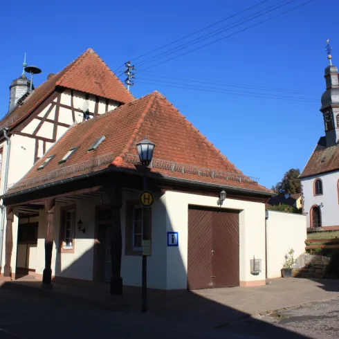 Ein traditionelles Fachwerkhaus mit einem roten Dach steht an einer ruhigen Straße. Im Hintergrund ist eine Kirche mit zwei Türmen zu sehen.