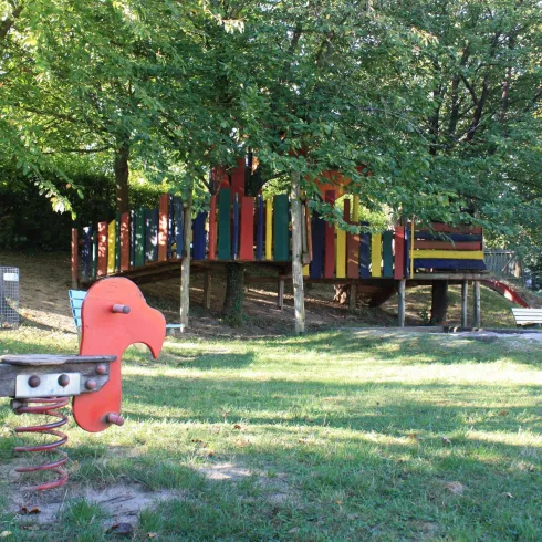 Ein bunter Spielplatz mit einer erhöhten Plattform und mehreren Baumgruppen. Im Vordergrund steht ein Federspielzeug in Form eines Vogels.