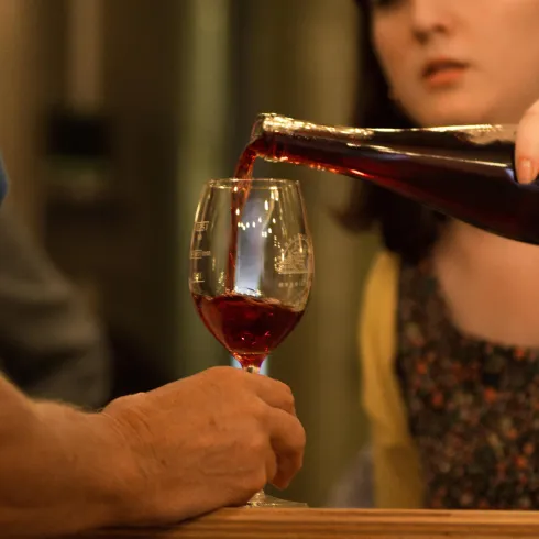 Eine Person schenkt Wein in ein Glas. Im Vordergrund ist ein Handgelenk zu sehen, während im Hintergrund eine andere Person schaut.