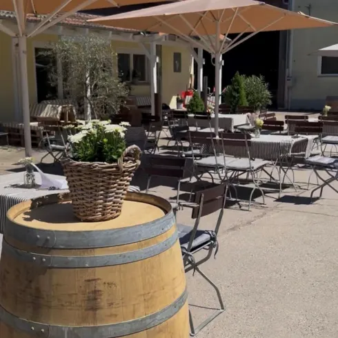 Eine gemütliche Terrasse mit Tischen und Stühlen unter Sonnenschirmen. Im Vordergrund steht ein Holzfass mit einer Blumenschale.