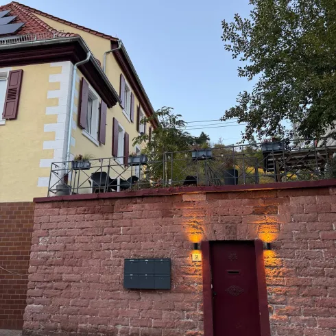 Ein charmantes Haus mit gelber Fassade und roten Fensterläden. Im Vordergrund ist eine Mauer mit einem Tor und sanfter Beleuchtung zu sehen.