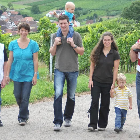 Eine Gruppe von sechs Personen spaziert in einem Weinberg. Im Hintergrund sind grüne Hügel und kleine Häuser zu sehen.