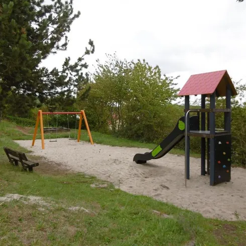 Ein kleiner Spielplatz mit einer Rutsche und einer Schaukel in einem sandigen Bereich. Im Hintergrund wachsen einige Bäume und Sträucher.