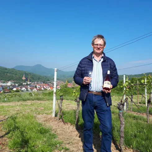 Ein Mann steht in einem Weinberg mit einer Flasche Wein in der Hand. Die Landschaft zeigt sanfte Hügel und einen klaren blauen Himmel im Hintergrund.