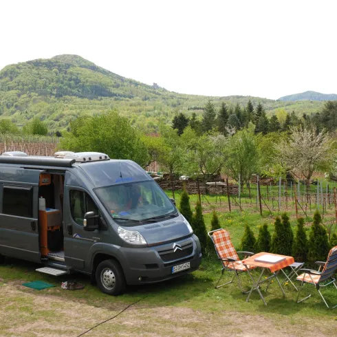 Ein grauer Camper steht ruhend in einer malerischen Landschaft mit sanften Hügeln und Weinreben. Neben dem Fahrzeug stehen zwei Stühle und ein Tisch auf einer grünen Wiese.