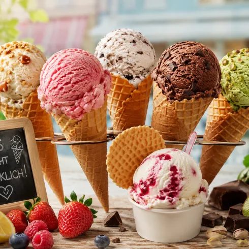 Sechs verschiedene Eiskugeln in Waffeln stehen auf einem Tisch. Neben den Eißorten liegen frische Früchte und eine Tafel mit der Aufschrift „EIS MACHT GLÜCKLICH!“.