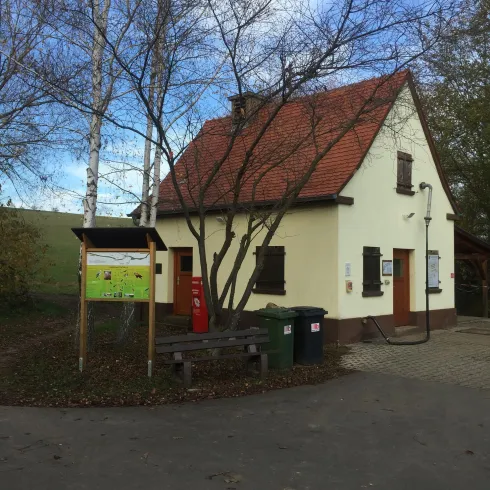 Ein kleines, zweigeschossiges Haus mit rotem Dach und mehreren Bäumen im Vordergrund. Neben dem Haus steht ein Informationsschild und es gibt Mülltonnen.