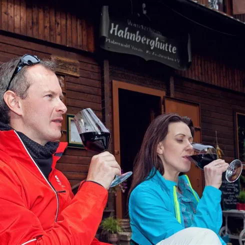 Ein Paar genießt Wein vor einer rustikalen Berghütte. Die beiden sind in Freizeitkleidung und entspannen in der Natur.