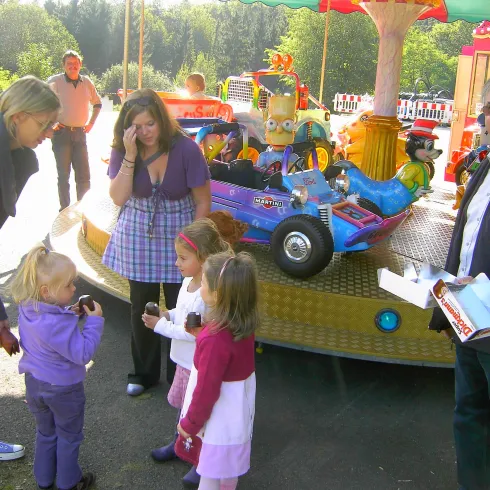 Ein bunter Platz mit einem Karussell und fröhlichen Kindern. Erwachsene stehen daneben und beobachten die Spielenden.