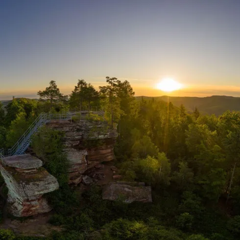 Ein schöner Aussichtspunkt auf einem Felsen umgeben von Bäumen. Im Hintergrund ist ein Sonnenuntergang über den Hügeln zu sehen.