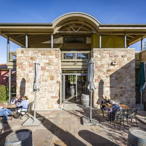 Ein modernes Gebäude mit Natursteinfassade und großen Fenstern. Auf der Terrasse sitzen Menschen, die die Sonne genießen.