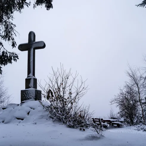Ein großes Kreuz steht inmitten einer schneebedeckten Landschaft. Die kühle, graue Atmosphäre und die kahlen Bäume verleihen dem Ort eine ruhige Stimmung.