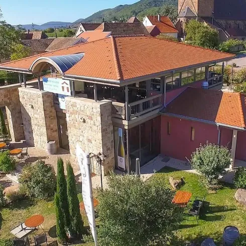 Ein modernes Gebäude mit roten Ziegeldächern und großen Fenstern, umgeben von einem gepflegten Garten. Es gibt Terrassen mit Tischen und Stühlen für Besucher.
