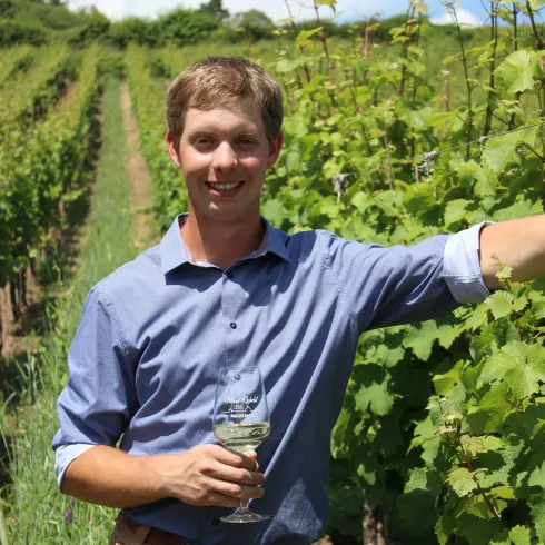 Ein junger Mann steht lächelnd in einem Weinberg und hält ein Glas Wein in der Hand. Die Reihen von Weintrauben sind grün und üppig, unter einem klaren blauen Himmel.