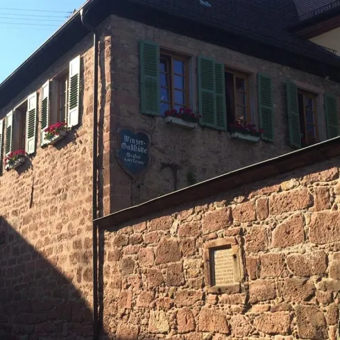 Ein historisches Gebäude mit einem steinernen Mauerwerk und Fensterläden. Bunte Blumen in Fensterkästen verleihen dem Ort Charme.