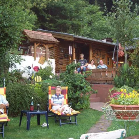 Eine gemütliche Gartenlandschaft mit einem Holzhaus im Hintergrund. Vier Menschen entspannen in Liegestühlen, umgeben von grünen Pflanzen und bunten Blumen.