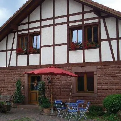 Ein charmantes Fachwerkhaus mit roten Akzenten und bunten Blumen in den Fenstern. Vor dem Haus stehen Tische und Stühle unter einem roten Sonnenschirm.