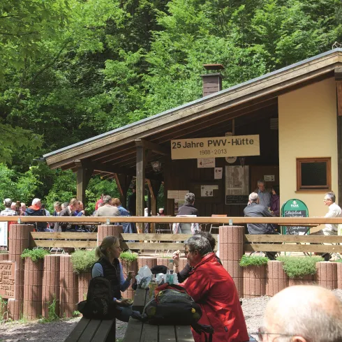 Eine Hütte im Wald mit einer Gruppe von Menschen, die sich versammelt haben. Die Umgebung ist grün und einladend.