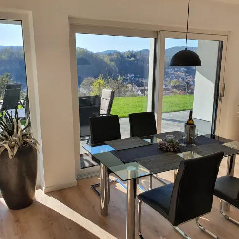 Ein eleganter Essbereich mit einem Glastisch und schwarzen Stühlen. Große Fenster bieten einen schönen Blick auf die Landschaft.