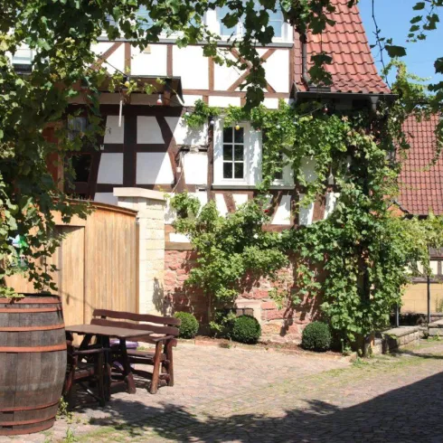 Ein malerischer Innenhof mit Weinranken und einer alten Holzfass. Im Hintergrund sind Fachwerkhäuser und eine gemütliche Sitzgelegenheit zu sehen.