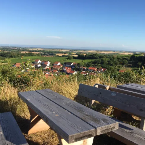 Eine Holzbank mit Tisch steht auf einem Hügel und bietet einen Blick auf ein malerisches Dorf und die umliegende Landschaft. Der Himmel ist klar und blau, und die Felder sind grün.
