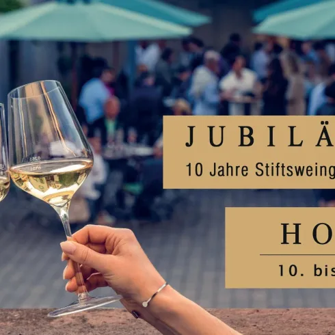 Zwei Gläser mit Weißwein stoßen an, um einen Jubiläum zu feiern. Im Hintergrund ist eine festliche Atmosphäre mit Menschen und grünen Sonnenschirmen zu sehen.