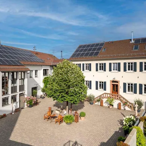 Ein charmantes Anwesen mit zwei Gebäuden und einem großen Baum im Innenhof. Das Dach des linken Gebäudes ist mit Solarpanelen ausgestattet.