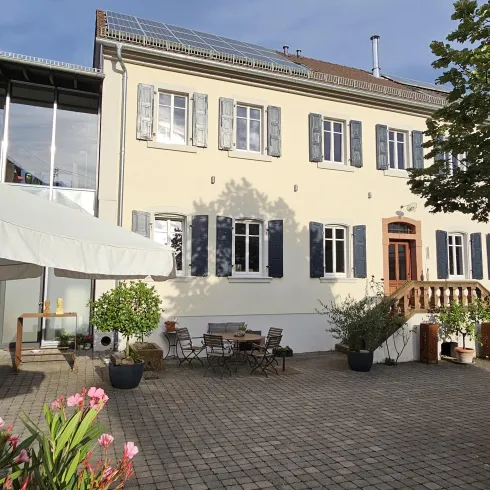 Ein elegantes Haus mit weißer Fassade und blauen Fensterläden. Der Außenbereich ist schön gestaltet mit Blumen und einer Sitzgelegenheit.