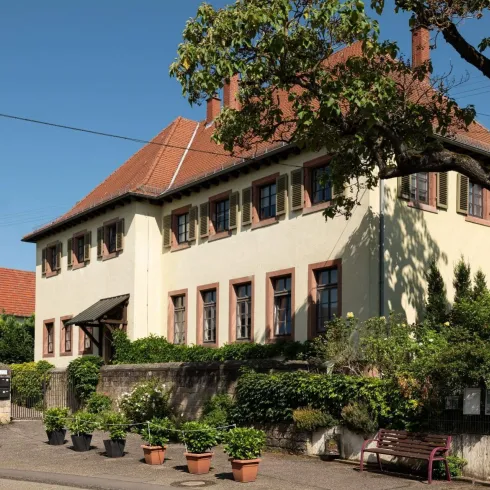 Ein großes, helles Gebäude mit einem roten Satteldach und vielen Fenstern. Umsäumt von grünen Pflanzen und Bäumen bietet es eine einladende Atmosphäre.