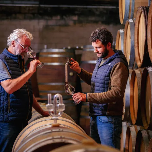 Zwei Männer stehen in einem Weinkeller neben Holzfässern. Sie prüfen den Wein und sprechen miteinander.