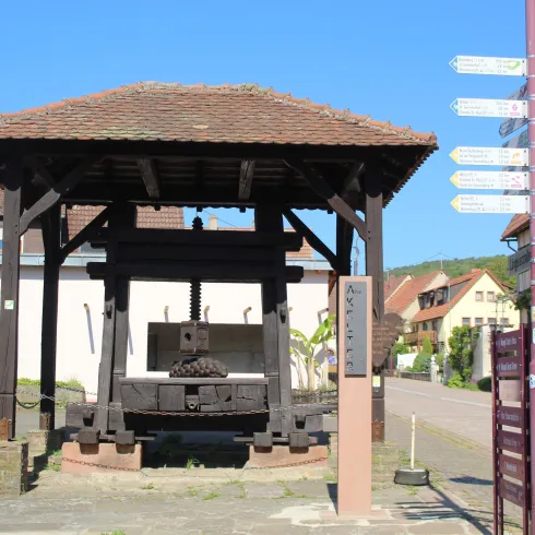 Ein traditioneller Holzunterstand mit einer alten Maschine in der Mitte. Im Hintergrund sind einige Gebäude und Wegweiser zu sehen.