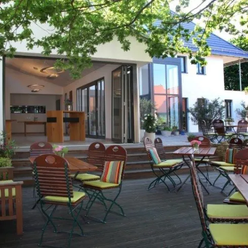 Ein gemütlicher Außenbereich mit Tischen und Stühlen unter einem Baum. Im Hintergrund ist ein modernes Gebäude mit großen Fenstern zu sehen.