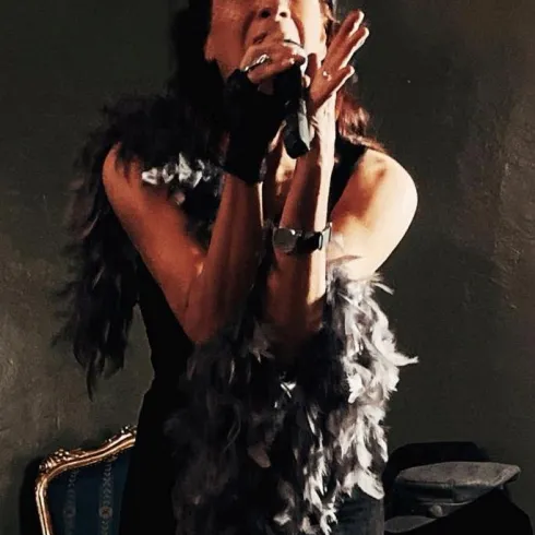 Eine Sängerin trägt ein schwarzes Outfit und einen Federboa. Sie performt leidenschaftlich auf der Bühne.