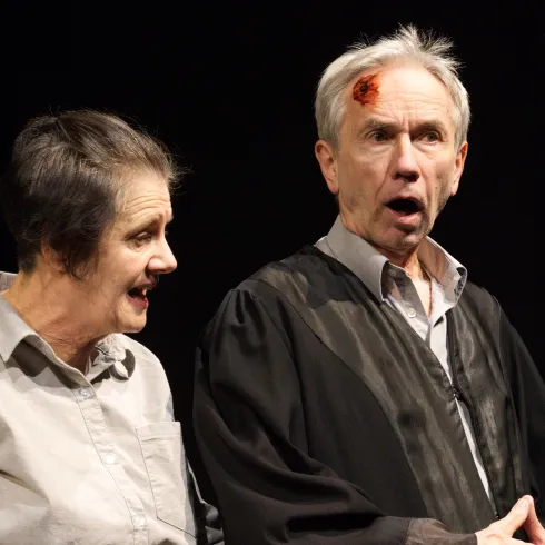 Zwei Schauspieler auf der Bühne in einem emotionalen Moment. Der Mann trägt eine Robe und hat eine Verletzung am Kopf, während die Frau ihn ansprechen scheint.