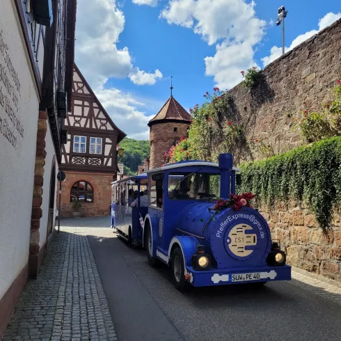 Ein blauer Touristenzug fährt durch eine malerische, enge Gasse. Im Hintergrund sind traditionelle Fachwerkhäuser und ein kleiner Turm sichtbar.