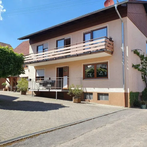 Ein einladendes Wohnhaus mit einem Balkon und einem gepflasterten Vorhof. Der Himmel ist klar und blau, was eine angenehme Atmosphäre schafft.