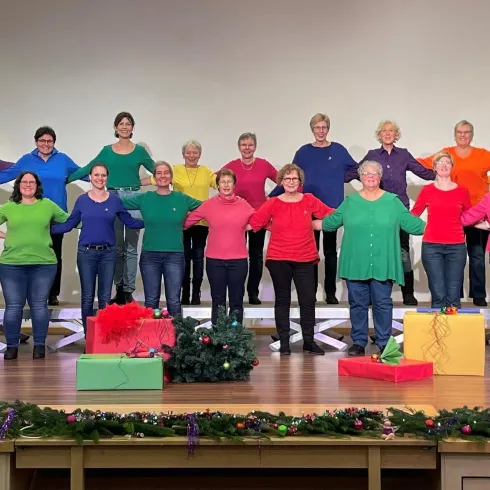 Eine große Gruppe von Menschen steht in bunten Oberteilen auf einer Bühne. Vor ihnen befinden sich Geschenke in verschiedenen Farben.