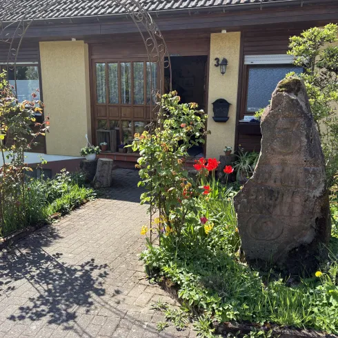 Ein gemütliches Haus mit einem einladenden Eingang und bunten Blumen im Garten. Ein markanter Stein neben dem Weg ergänzt die idyllische Atmosphäre.