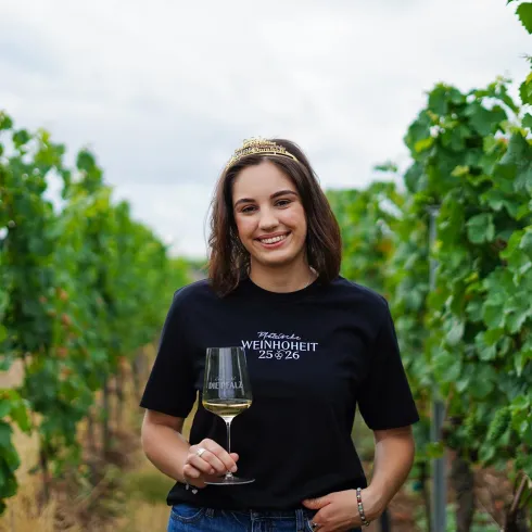 Eine Frau in einem Weingarten hält ein Glas Wein in der Hand. Sie trägt ein schwarzes T-Shirt und lächelt vor einer Reihe von Weinreben.