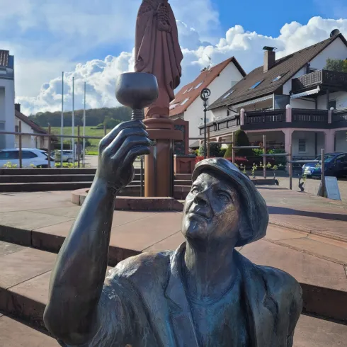 Eine bronze Statue eines Mannes, der einen Kelch hält, steht im Vordergrund. Im Hintergrund ist eine weitere Statue sowie charakteristische Gebäude zu sehen.